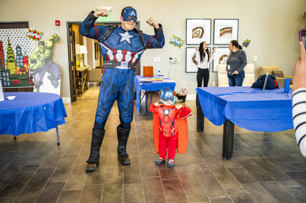 Adult and child dressed as captain america flexing muscles at a party with people watching, in a room decorated with blue tablecloths and artwork on the walls.