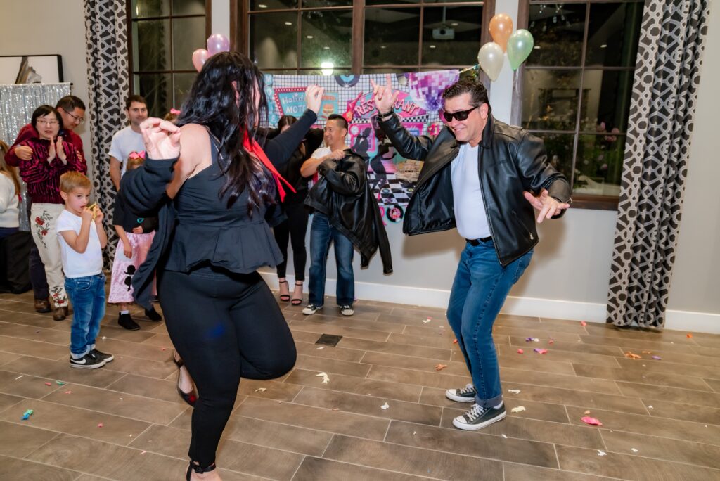 A lively party scene with adults and children. two people in the foreground are dancing energetically, one dressed in a black leather jacket and sunglasses. balloons and festive decorations in the background.