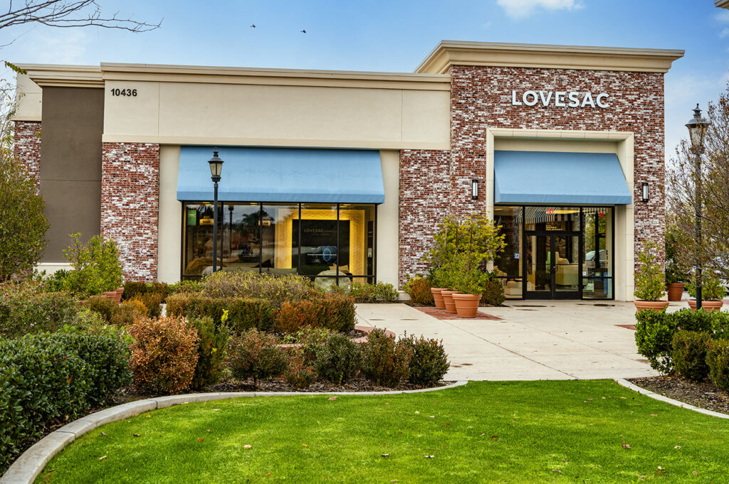 Exterior view of a lovesac store with a distinctive brick facade and large windows, featuring blue awnings and landscaped garden in front. a lamp post and flying birds are also visible.