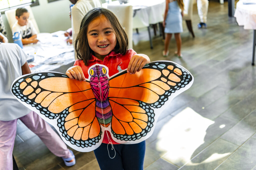 A smiling young girl is indoors, holding a large, brightly colored paper butterfly. The butterfly has vibrant orange wings with black and white patterns. Several other children are present in the background, engaged in activities at various tables.