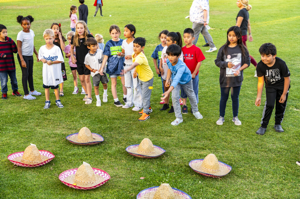 A group of diverse children in casual clothes stands on a grassy field, looking excitedly at sombreros placed on the ground in front of them, preparing for a game.