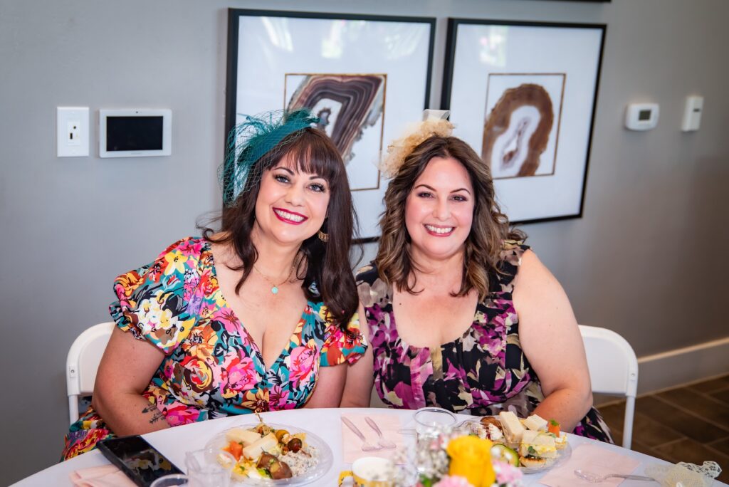 Two women are seated at a table, smiling at the camera. They are dressed in colorful floral dresses and wearing decorative headpieces. The table has plates of food and floral centerpieces. The background features two abstract framed artworks on a gray wall.