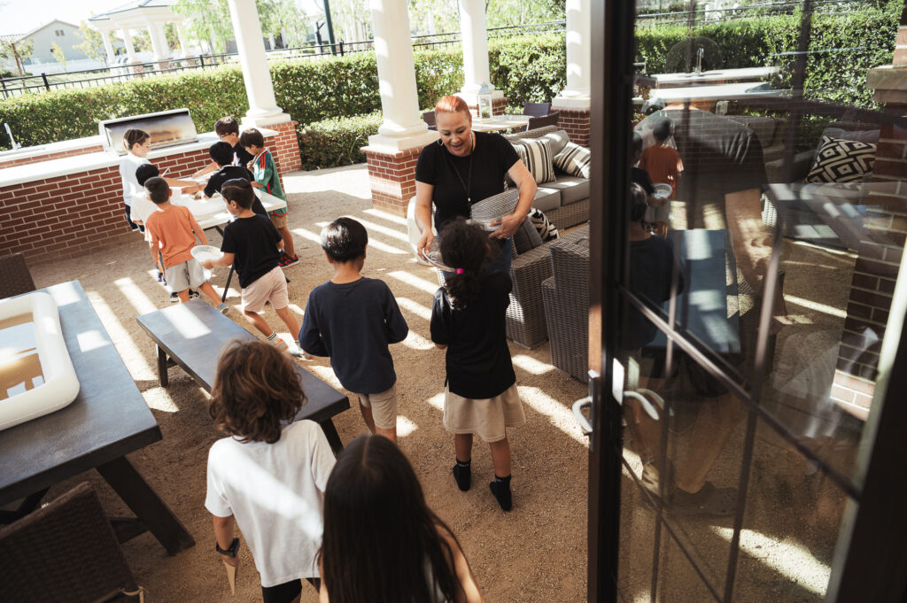 A group of children is gathered on a covered patio, engaged in an activity with a smiling adult. The space is furnished with tables, chairs, and a couch. Sunlight filters through the trees and the open windows, creating a bright and lively atmosphere.