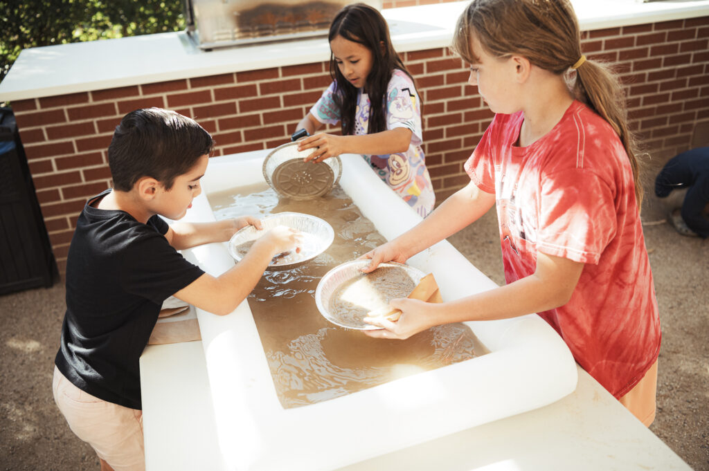 Three children are engaged in an outdoor activity, sifting gravel and sand in a water-filled table. They are focused on the task, using round pans to search for items, with a brick wall in the background.