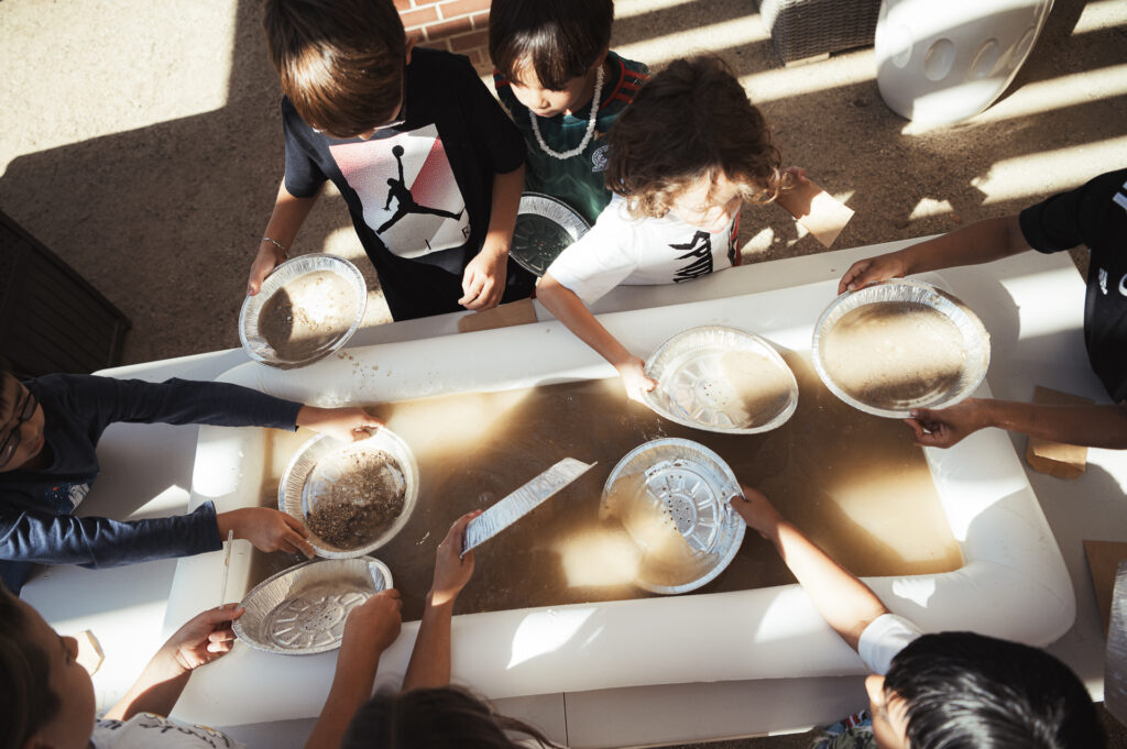 Children play with water and mud using aluminum pie pans at a long rectangular table outdoors. The sunlight casts shadows across the scene.