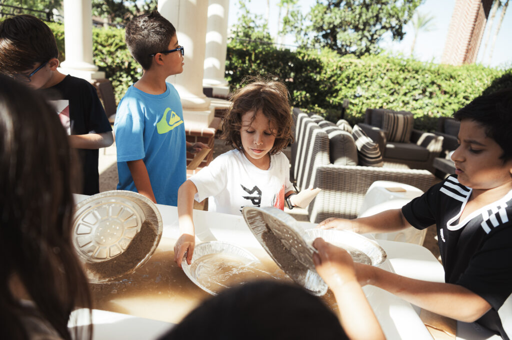 A group of children stand around a water table outdoors, using metal pie tins. They appear focused and engaged, with sunlight filtering through nearby trees and columns. Patio furniture is visible in the background.