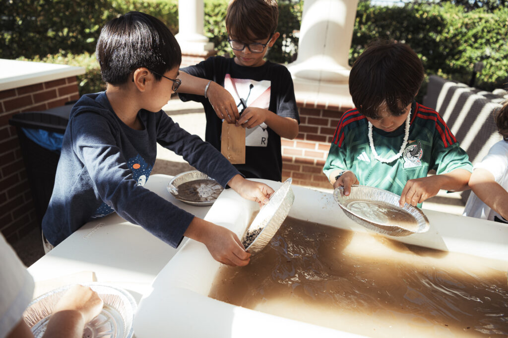 Children are participating in a hands-on activity involving water and pans at an outdoor table. They appear to be panning for objects or materials. The scene is set in a sunny area with trees and brick columns in the background.