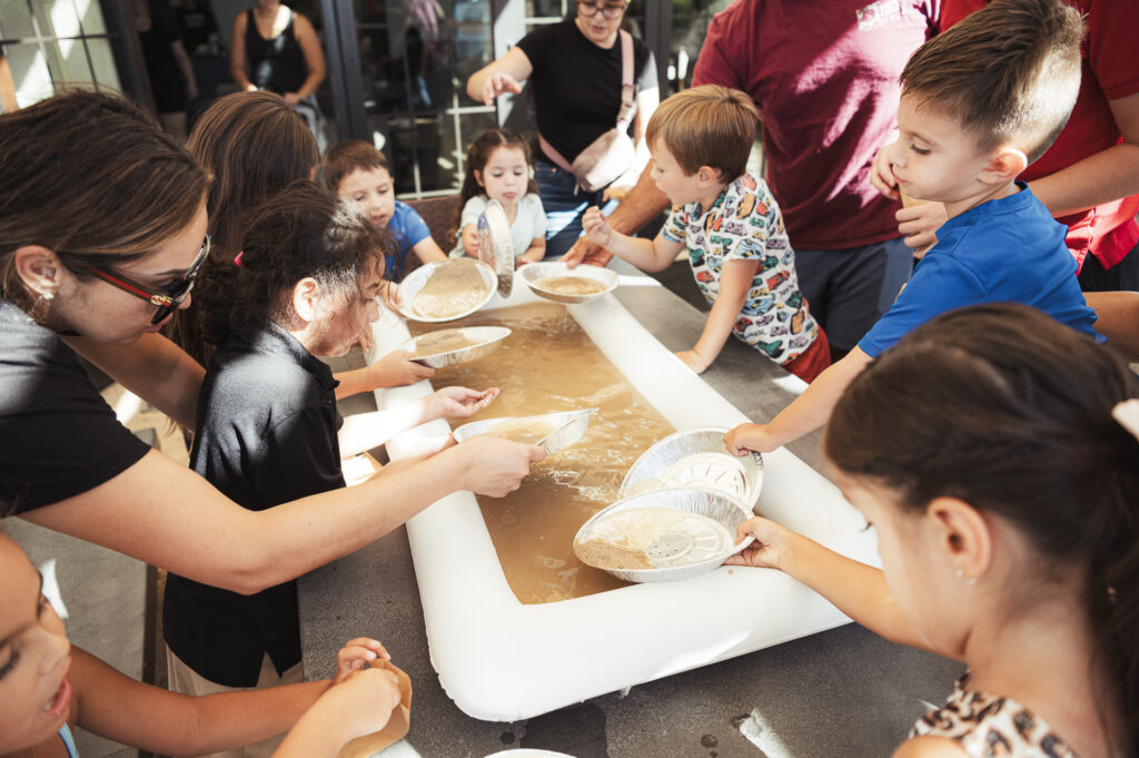 Children and adults gather around a long table engaging in an activity that involves sifting through water with pans, possibly for a fun treasure or panning activity. The group is focused and interacting enthusiastically.