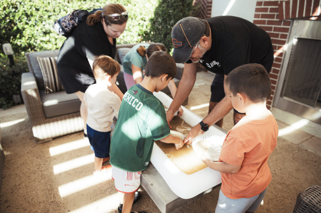 A group of children and two adults are gathered around a large, white outdoor table, engaged in a hands-on activity. The children are focused, and the setting is a backyard patio with comfortable seating.