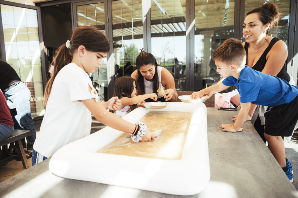 Children and adults gather around a table with a shallow inflatable tray filled with sand and small shovels. They are engaged in a fun, hands-on activity. Natural light from the building's large windows brightens the scene.