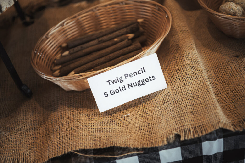 A wicker basket filled with twig-shaped pencils is placed on a burlap cloth. A sign in front reads "Twig Pencil 5 Gold Nuggets." The tablecloth beneath is checkered black and white.