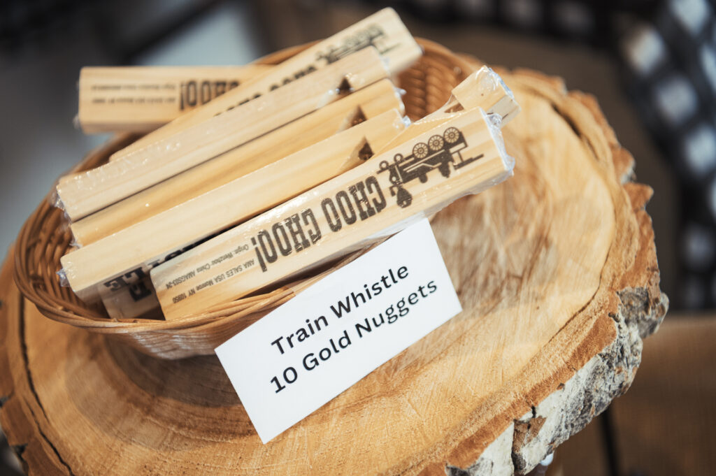 A basket of wooden train whistles is displayed on a tree stump slice. A sign in front reads "Train Whistle 10 Gold Nuggets." The whistles have train illustrations and text printed on them.