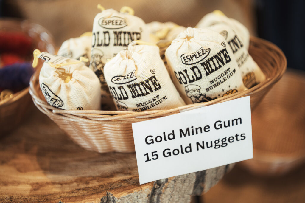 A woven basket holds several small fabric sacks labeled "Gold Mine Nugget Bubble Gum." A sign in front of the basket reads "Gold Mine Gum, 15 Gold Nuggets." The setting suggests a rustic or vintage theme.