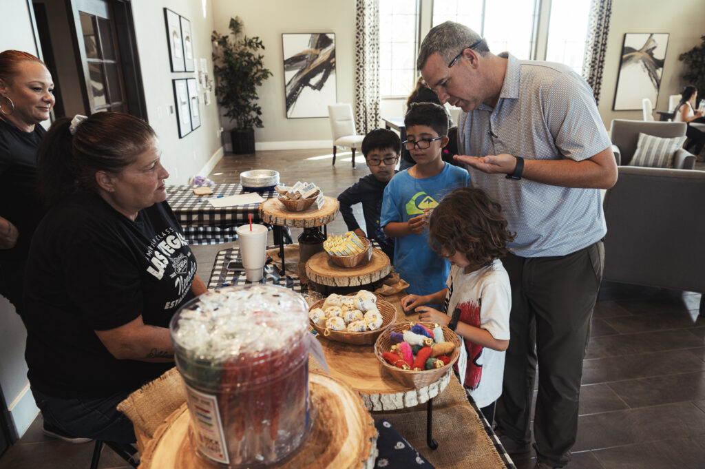 A group of people, including children and adults, gather around a table with various pastries and treats on display. One child is reaching for snacks, while others are interacting. The setting is a cozy indoor space with natural light coming through the windows.