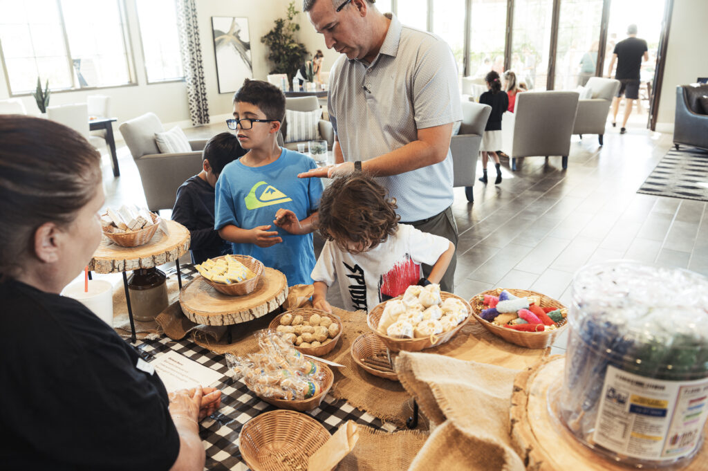 A family gathers at a table filled with various baked goods and candies. A man and two boys browse the selection, while a woman assists them. The room has large windows and several people seated in the background.