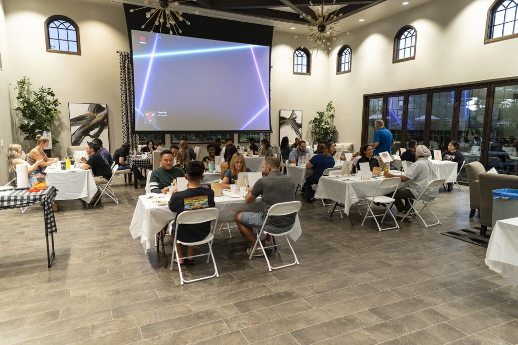 A group of people seated at tables in a large room with white tablecloths. A large screen displays a colorful triangle pattern. The room has high ceilings, large windows, and indoor plants. The atmosphere is casual and social.