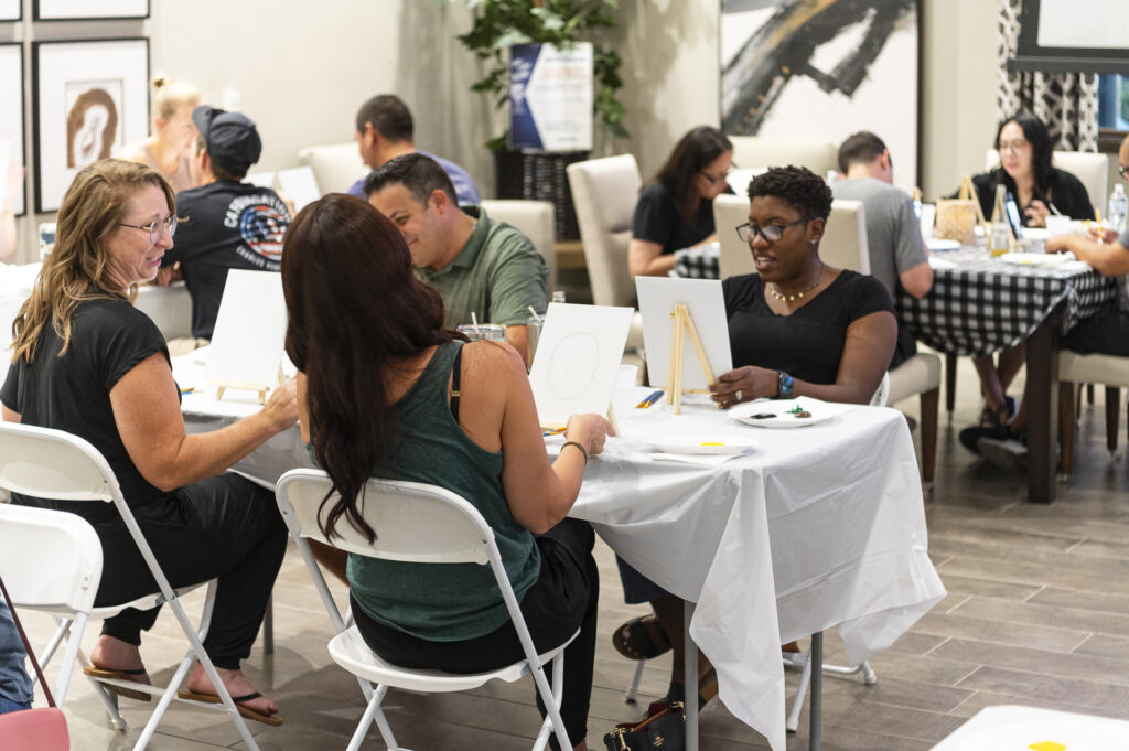 People are seated around tables in a bright room, engaged in a painting workshop. They are focused on easels with blank canvases, while chatting and exchanging ideas. The atmosphere is lively and creative, with art supplies and decorative artwork visible.