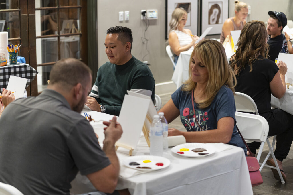 People seated at tables participate in a painting class. Each person has a canvas and paint supplies in front of them. The room has a casual and creative atmosphere, with participants focused on their art projects.