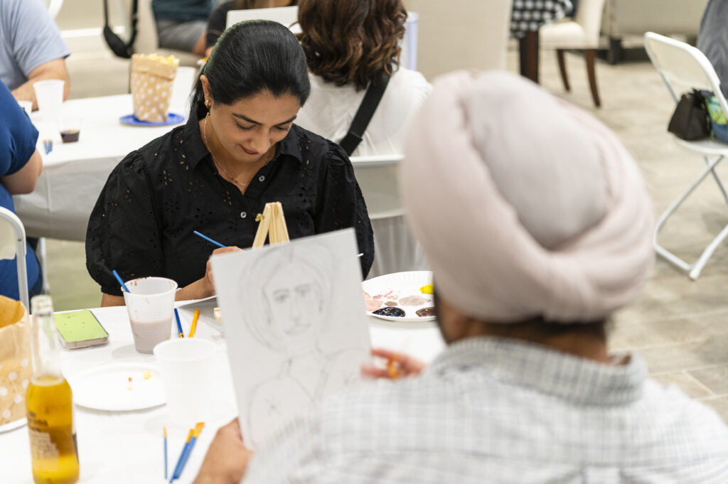 A woman seated at a table is painting on a small canvas. In the foreground, a person wearing a turban is drawing with a pencil. The table is scattered with art supplies, cups, and a bottle. The background shows more people and furniture.