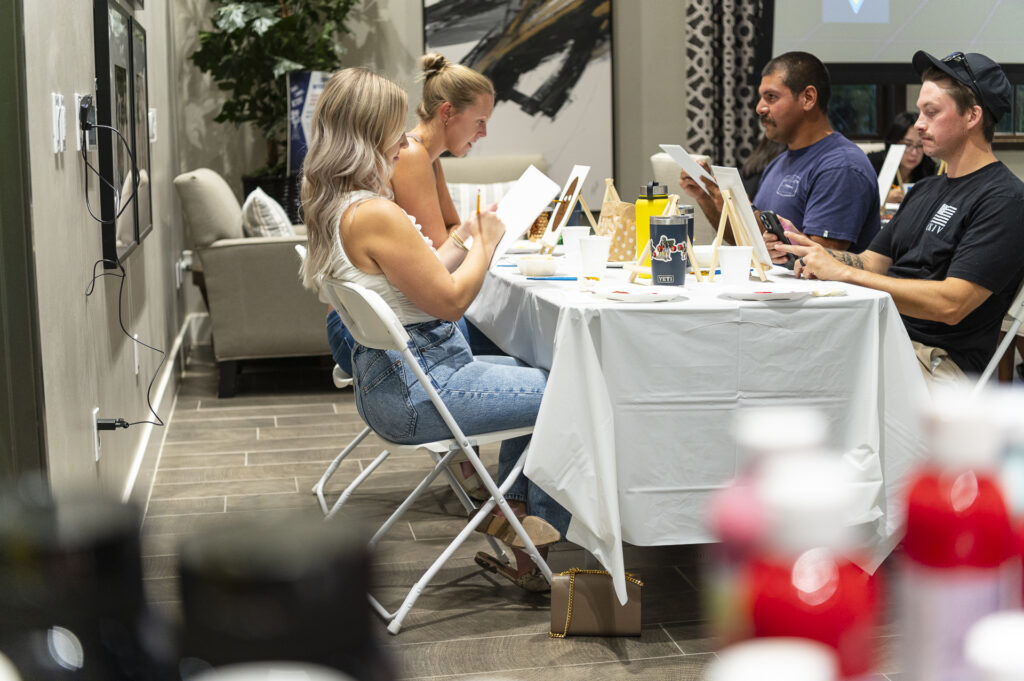 A group of people sit at a table engaged in painting activities. The room is bright, with canvases and art supplies scattered on the table. A large painting and potted plant are visible in the background.