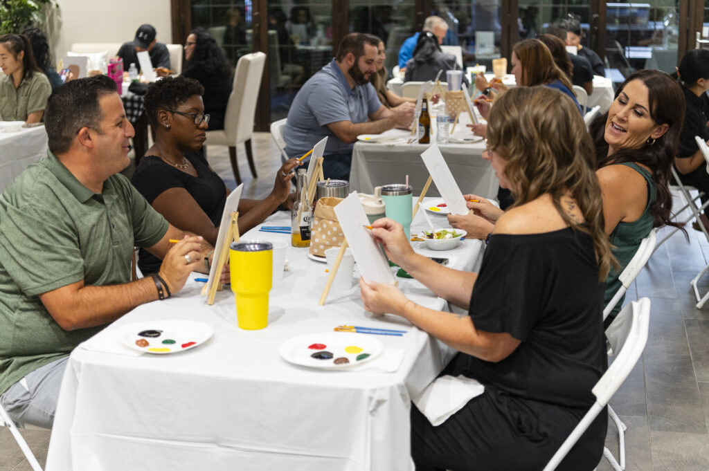 People seated at tables engaged in a painting activity, with canvases, paint, and brushes. The atmosphere is relaxed and social, with participants smiling and chatting.