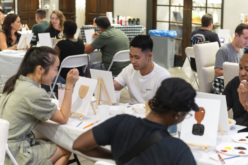 People are seated at tables participating in a painting class, with canvases, paints, and brushes in use. The room has a casual atmosphere, with participants focused on their artwork around white plastic-covered tables.