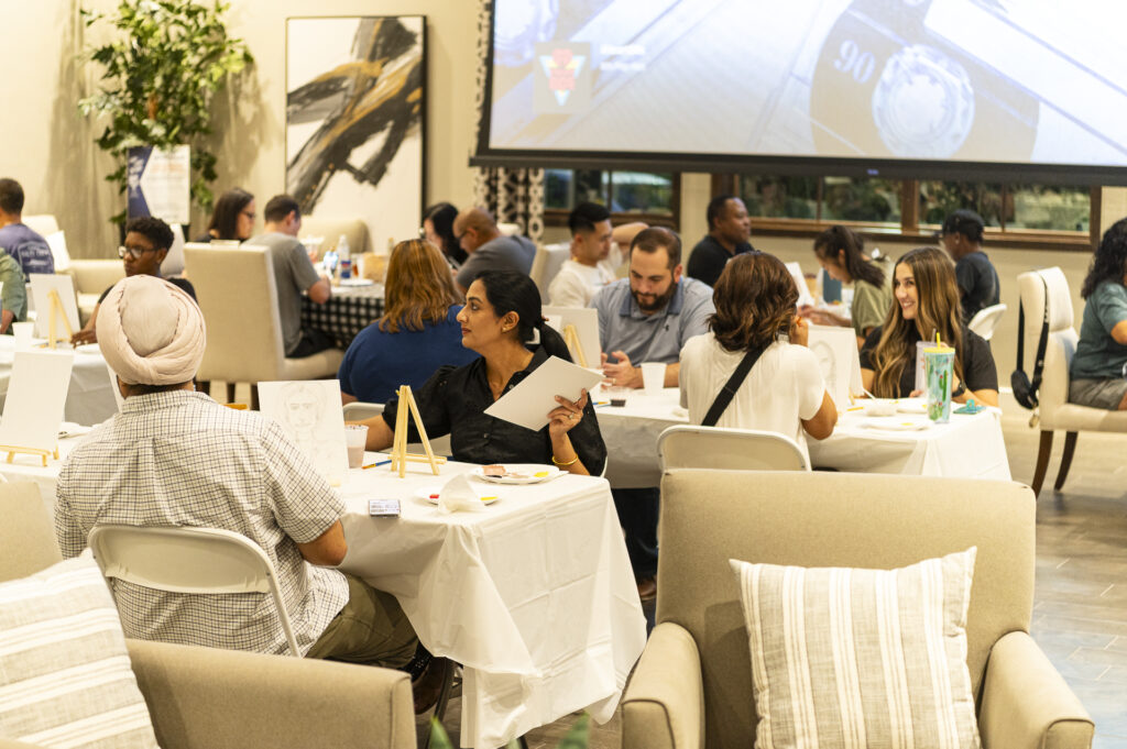 A group of people seated at tables covered with white cloths, engaged in an art activity. Some individuals are painting on canvases. The room has a large screen at the front, plants, and modern decor. People are chatting and focusing on their projects.