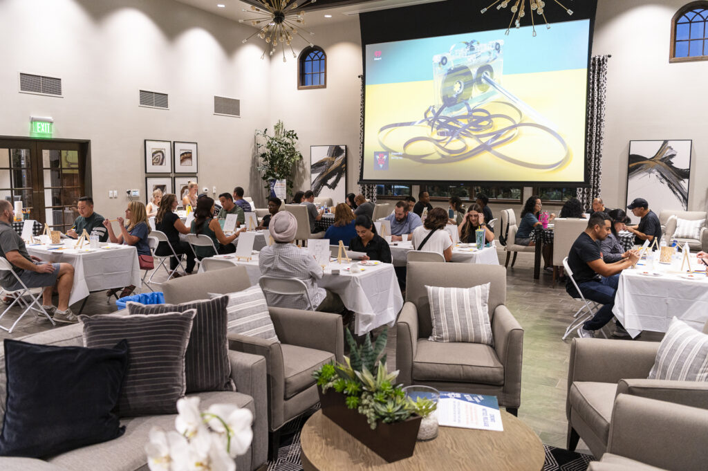 A group of people seated at tables in a well-lit room with beige walls and high ceilings. There is a large screen displaying an image of a device with cables. Comfortable seating includes sofas and chairs. Various art pieces and plants decorate the space.