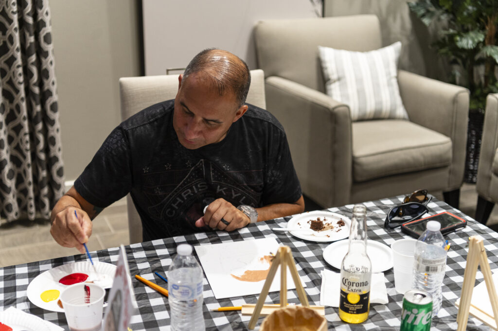 A person is painting at a table covered with a black and white checkered tablecloth. Art supplies, drinks, and a slice of cake are on the table. Comfortable chairs and a curtain are in the background, creating a relaxed atmosphere.