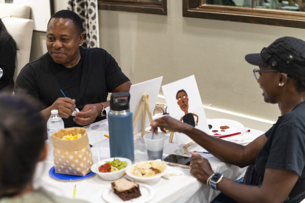 Two people sit at a table, painting and chatting. The table holds snacks, art supplies, and a portrait. Both individuals appear engaged and smiling, suggesting a relaxed, creative atmosphere.