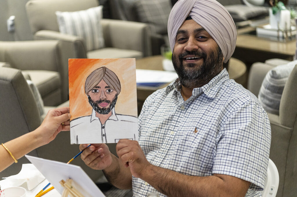 A man wearing a turban and checkered shirt smiles while holding up a drawing of himself. The artwork features a similar portrayal with an orange background. They are seated in a room with comfortable chairs.