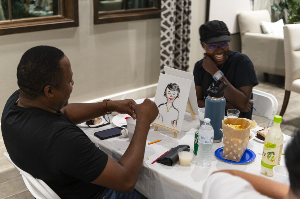 Two people sitting at a table, smiling and enjoying a painting session. One person holds a brush, working on a canvas with a sketch of a person. The table has art supplies, drinks, and a popcorn container. A cozy room setting surrounds them.