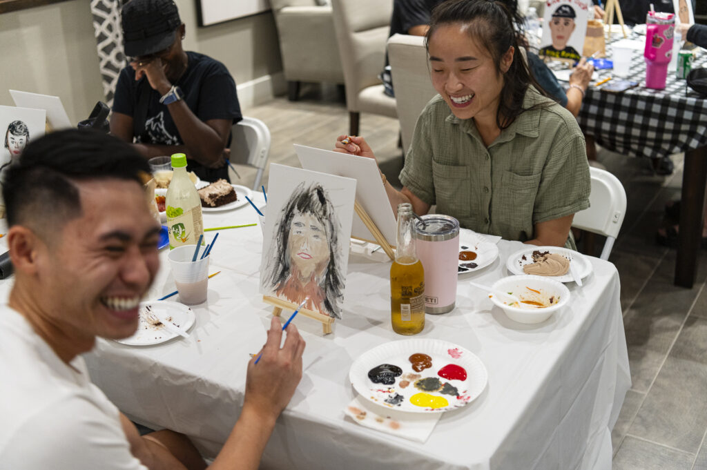 People are sitting at a table enjoying a painting activity. A man and a woman are laughing and painting on canvases. Paint supplies and beverages are on the table. The mood is cheerful and relaxed.