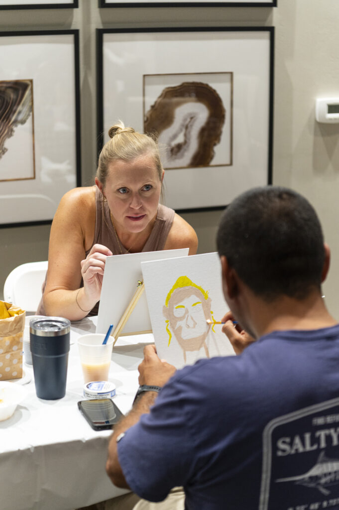 A woman attentively watches a man painting her portrait at a table. She sits with a drink and snack nearby. The man focuses on the canvas, wearing a t-shirt. They are surrounded by framed artwork on the wall.