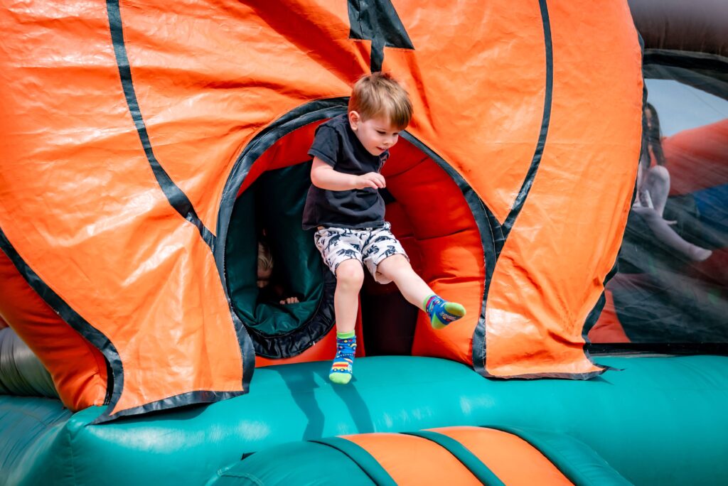 A young boy jumps out of a large inflatable structure, wearing a black shirt and patterned shorts. The inflatable has a bright orange and green design. The scene is outdoors and the child appears excited.