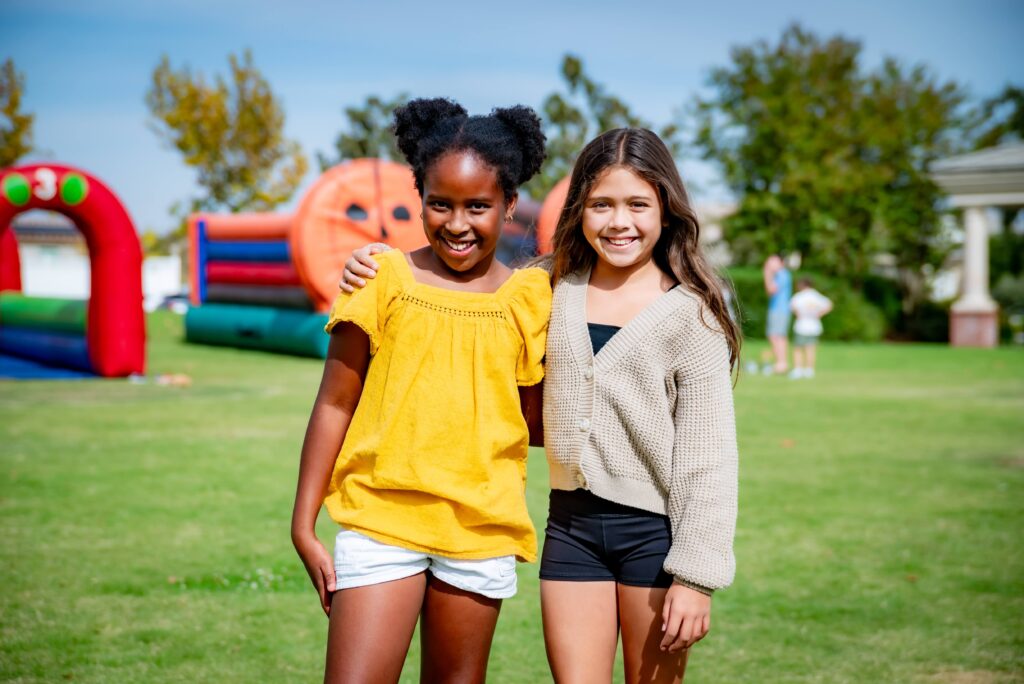 Two girls smiling and standing together on a grassy field with inflatable play structures in the background. One wears a yellow top and white shorts, the other a beige sweater and black shorts. It's a sunny day, and they appear to be at an outdoor event.