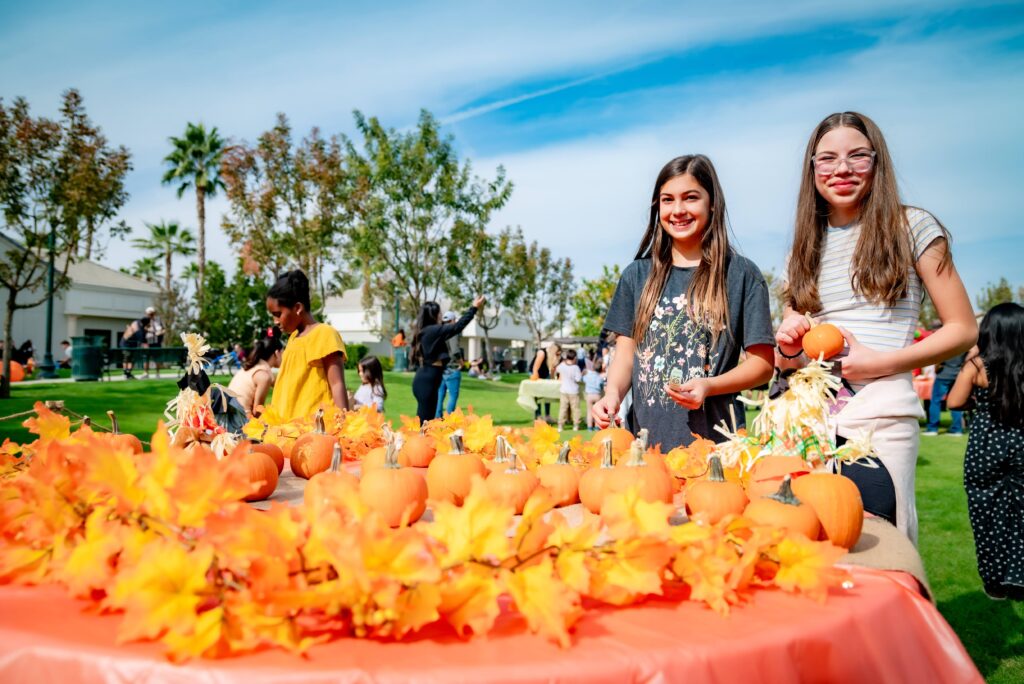 Two young girls stand beside a table decorated with small pumpkins and autumn leaves. The scene is set outdoors with trees and people in the background, under a sunny blue sky. The girls are smiling and holding craft materials.