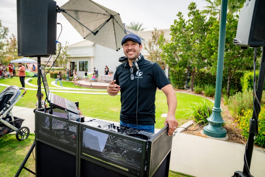 A DJ stands behind a console outdoors, wearing headphones around his neck and holding a phone. He smiles at the camera, surrounded by greenery. A stroller and a small crowd are visible in the background.