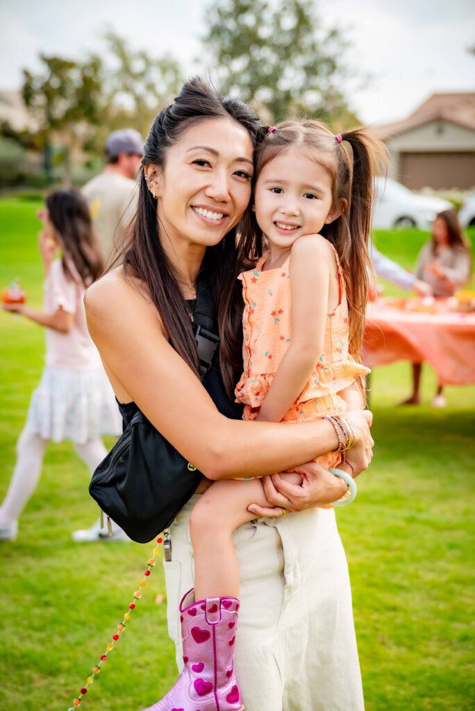 A woman smiles and holds a young girl in her arms at an outdoor gathering. The girl is wearing an orange top and pink heart-patterned boots. People are gathered around tables in the background on a grassy area.