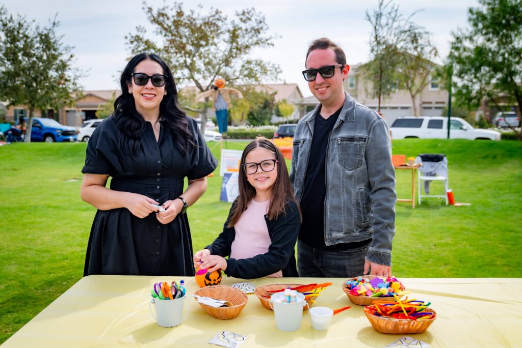 A family of three stands outdoors by a table with crafting supplies. The table displays bowls filled with colorful materials. Green grass and trees are in the background. All wear sunglasses and smile at the camera, enjoying a sunny day.