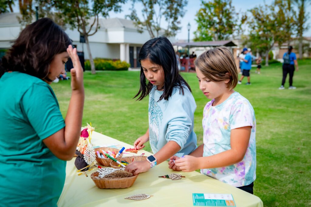 Three children stand at a table outdoors, engaging in a craft activity with baskets, leaves, and art supplies. They are focused and surrounded by a grassy lawn and trees, with buildings and other people in the background.