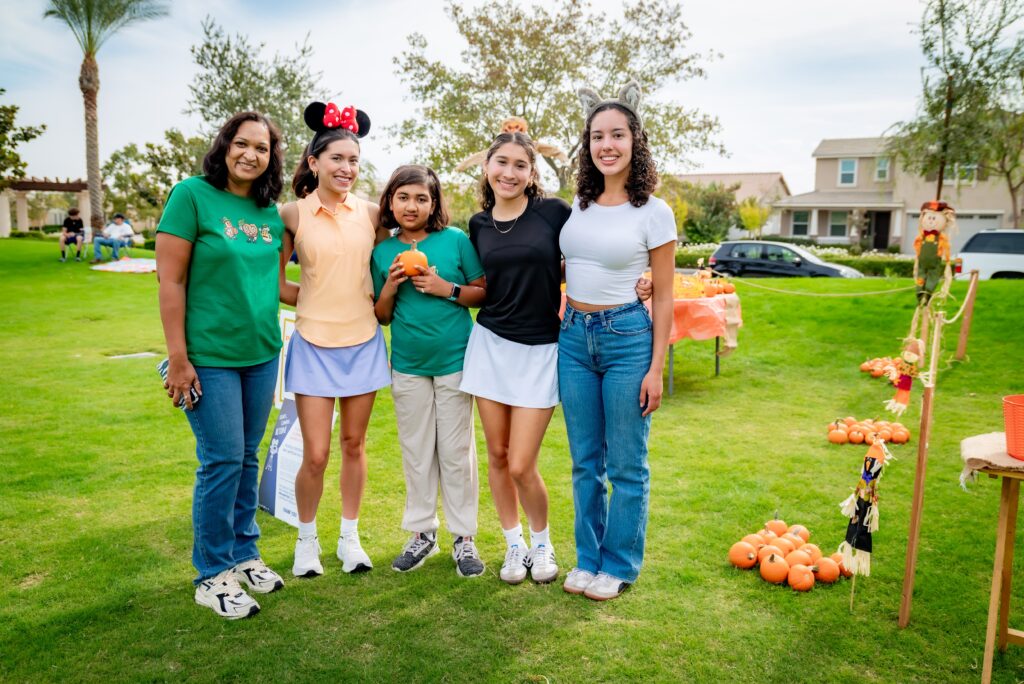 A group of five people stand on a grassy lawn, smiling at the camera. They are casually dressed, with two women wearing headbands. In the background, there's a table with pumpkins and trees.