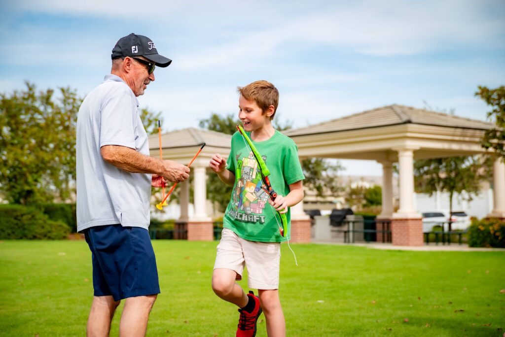 A man and a boy are outdoors on a grassy field, smiling and holding bow and arrow toys. The man is wearing a cap and sunglasses, while the boy is dressed in a green T-shirt and shorts. There are structures and trees in the background.