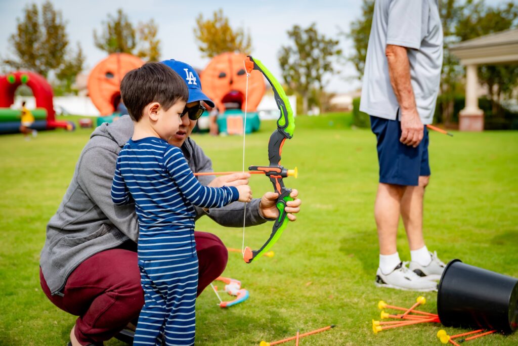 A man kneels beside a child holding a toy bow and arrow, guiding him on how to use it. They're outdoors on a grassy area with inflatable structures in the background. Another adult stands nearby, and toy arrows are scattered on the ground.