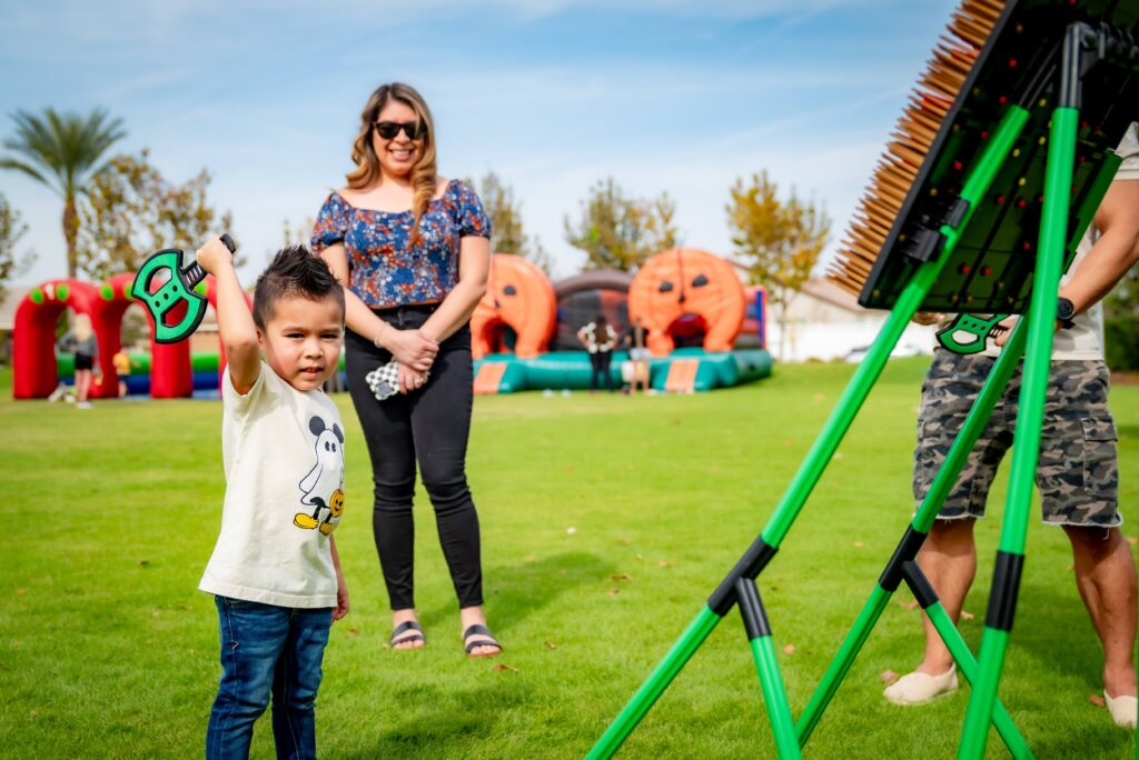 A young child with a toy hammer plays a game outside on a grassy field, standing near a large colorful booth. An adult watches in the background, smiling. Inflatable structures and clear blue skies are visible in the distance.