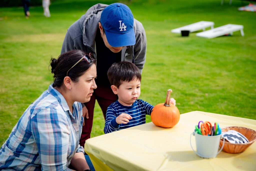 A young child, supervised by two adults, examines a small pumpkin on a table covered with a yellow cloth. Art supplies, including markers in a cup, are on the table. They are outdoors on a grassy field.