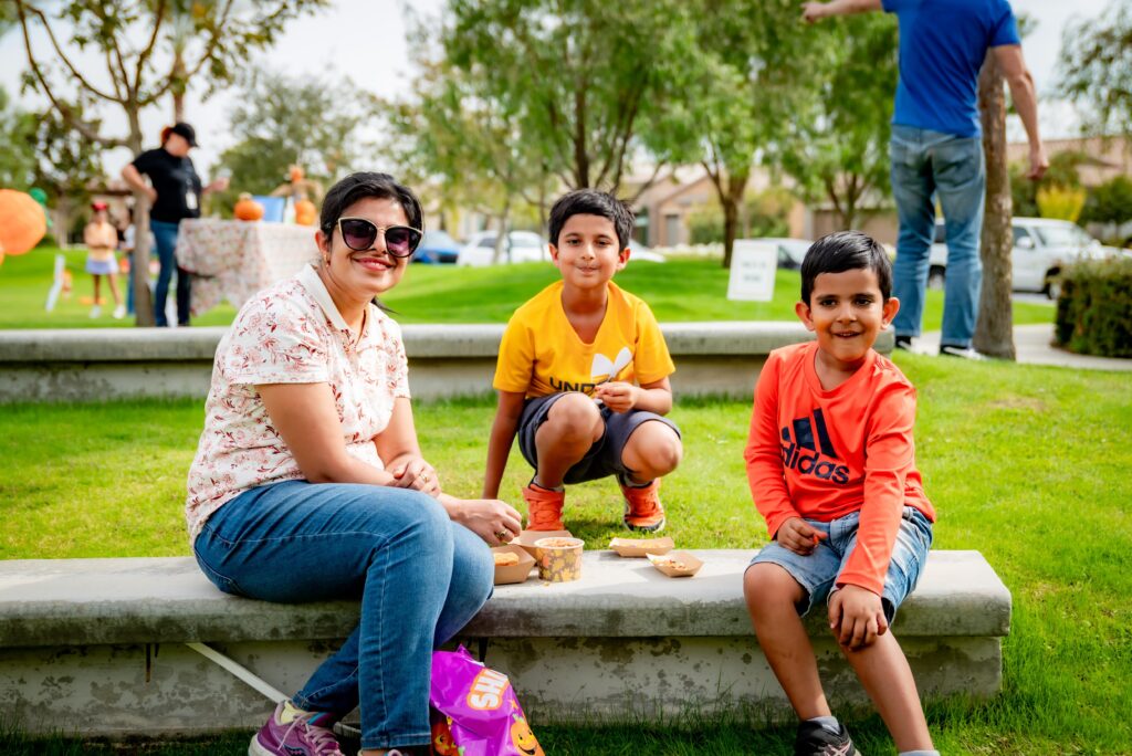 A woman and two young boys sit on a concrete bench in a park. The woman wears sunglasses and a floral shirt. The boys are in orange and yellow shirts. They are smiling and appear to be enjoying a sunny day. Others can be seen in the background.