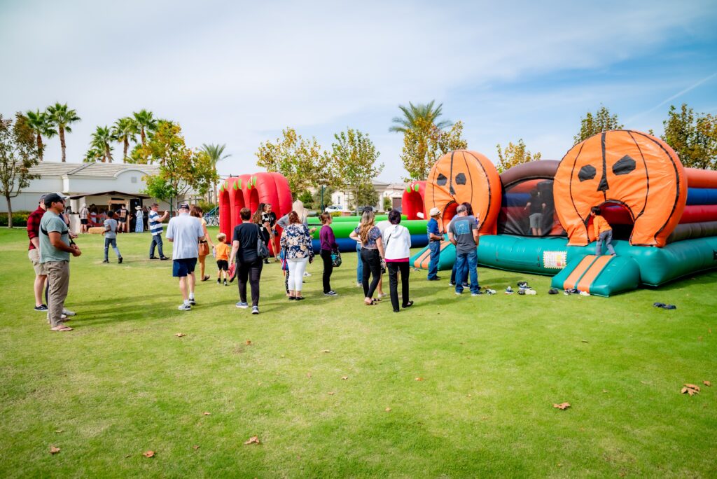 A group of people gathered around inflatable bounce houses shaped like pumpkins on a grassy field. The sky is clear with palm trees and other trees lining the background. Children and adults are enjoying the outdoor event.