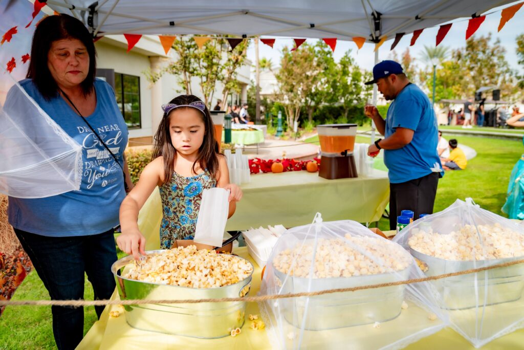 A girl is scooping popcorn into a bag at a stand under a canopy. Two large bowls of popcorn are on the table. A woman stands nearby, and another person is in the background near a beverage dispenser. Colorful banners decorate the canopy.