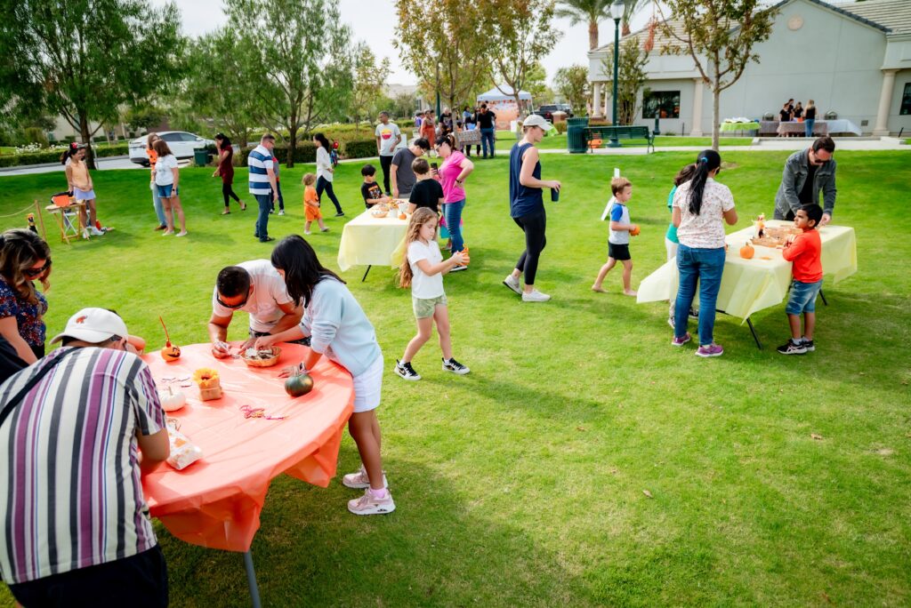 A community gathering in a park features people of various ages engaging in outdoor activities. Several tables are set up with crafts and pumpkins, and families and friends can be seen enjoying the event under a clear sky.